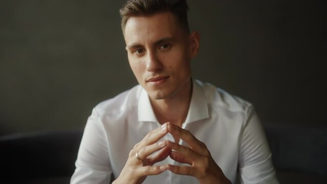 Confident young man in a white shirt, sitting with hands clasped, exuding calm professionalism and confidence. Portrait of handsome man in low key light against dark black solid background