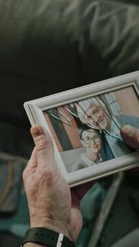 Vertical high angle shot of unhappy mature Caucasian couple separating after many years together. Wife packing suitcase and man looking at broken picture frame of them smiling at camera