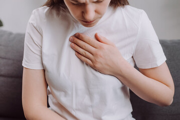 Unhealthy young woman sit on sofa having difficulty breathing pain of heart, touches his chest with hand. Trouble breathing, chest pain. Heart attack, thoracic osteochondrosis, panic attack concept