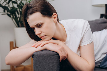 Close up of pensive stressed young caucasian woman sitting alone on sofa at home, looking away, thinking over problems, concern, touching face, feeling tired, concerned, hurt, headache concept