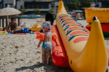 Dubai,  Unite dArab Emirates - October 19, 2019 - A child wearing arm floaties and a sun hat stands beside an inflatable banana boat on the beach, looking away from the camera.
