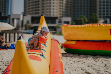 Dubai,  Unite dArab Emirates - October 19, 2019 - A small child with a sun hat and floaties sits on a large inflatable banana boat on the sand, focused on something in their hands.