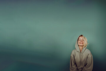 jeune femme blonde de face, avec des lunettes de vue et portant un sweater à capuche gris, rigolant, personne jeune et heureuse sur un fond vert dégradé avec espace négatif copy space - obrazy, fototapety, plakaty