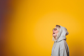 jeune femme blonde de profil, avec des lunettes de vue et portant un sweater à capuche gris, rigolant, personne jeune et heureuse sur un fond jaune moutarde dégradé avec espace négatif copy space - obrazy, fototapety, plakaty