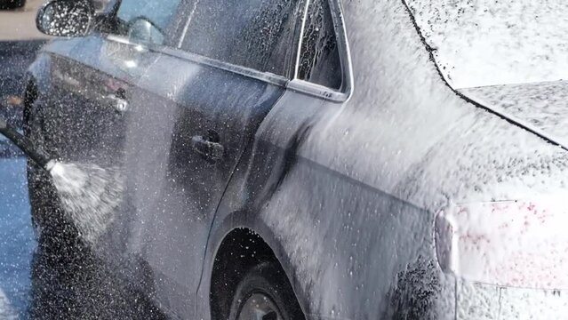 A vehicle is engulfed in soapy foam during a car wash, covering the tires, wheels, hood, and entire exterior of the car. Self-service car wash