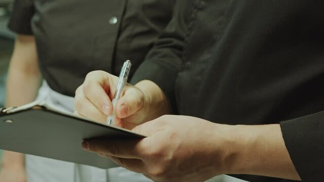 Head chef taking notes on clipboard and explaining job tasks to young female apprentice during workday in restaurant kitchen. Close-up, handheld shot