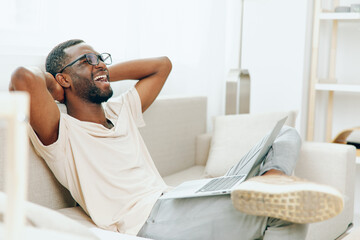 Young African American freelancer working on a laptop while sitting on a modern sofa in his cozy...