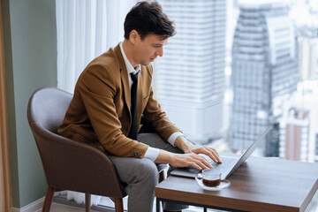 Portrait of skilled businessman working with laptop near window with skyscraper. Professional...