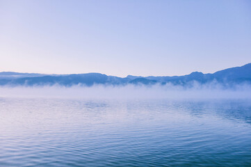 Picturesque scenery of the Lugu Lake in a thick white fog in sunrise. Silhouettes of mountains. Fall season. Nature, ecology, environmental conservation, eco tourism.