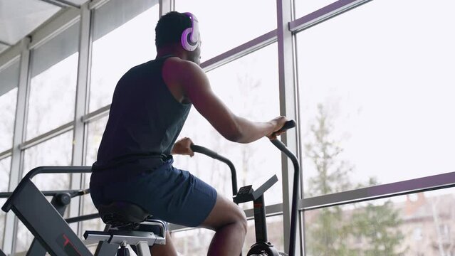 African american man with headphones doing cardio training on stationary air bike machine at the gym