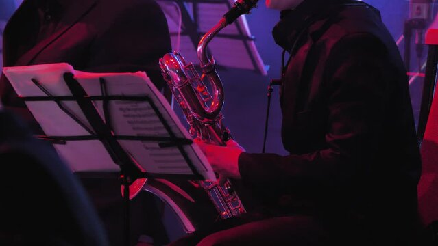 A musician in formal attire plays a baritone saxophone on a lit stage. A music stand with music sheets is visible. The atmosphere is dramatic and focused, emphasising the musician and his skill