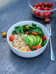 Bowl of food with brown rice, avocado, tomatoes, spinach, and a fork on a table