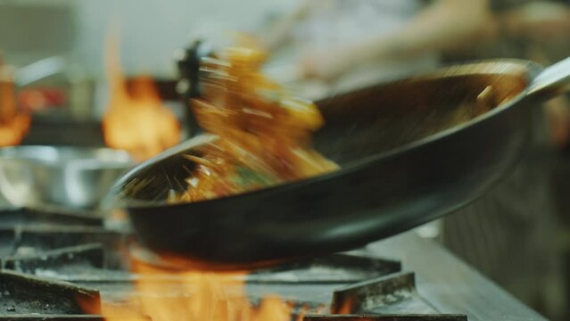 Close-up view of hands of master chef flipping food in frying pan as cooking on gas stove with burning fire flames in restaurant kitchen