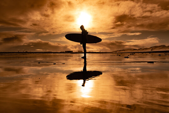 Woman standing on beach with surf board winter iceland sunset