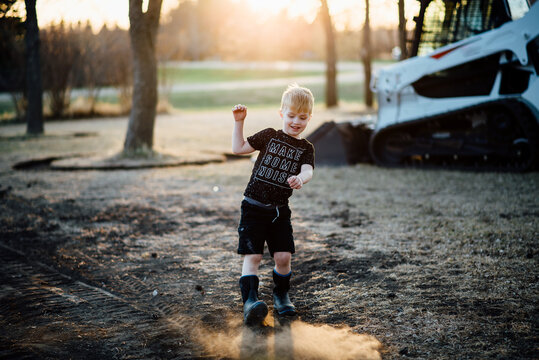 Wide view of young boy kicking up dirt in yard in front of skidsteer