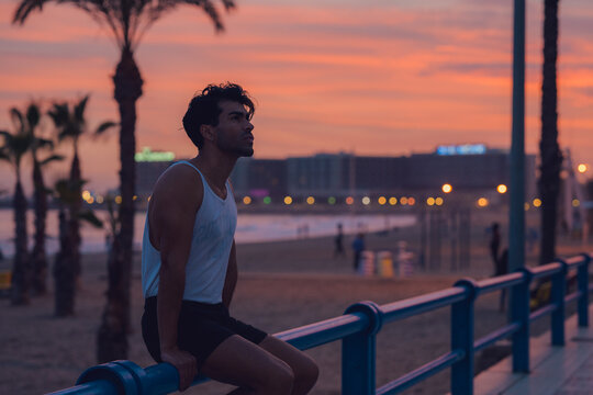 Pensive male athlete resting on a blue railing at beachside at sunset