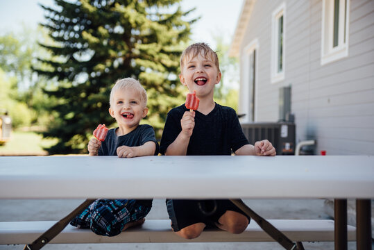 Two boys sitting on picnic table making silly faces with popsicles.