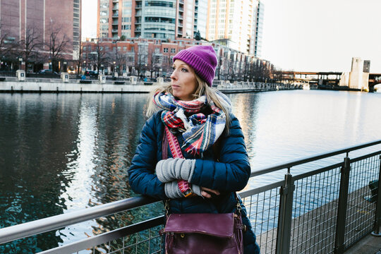 Female photographer in winter coat by river in city with camera bag