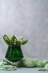 Keuken spatwand met foto White flowers, lilies of the valley in a green transparent vase, a glass, next to it lies a green scarf and three green balls. Gray background. Plenty of space for text © Alexander