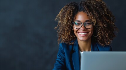 A businesswoman in a blue suit smiling while working on a laptop at the office. Portrait of a happy middle-aged woman with curly hair wearing glasses using a computer for work or online learning