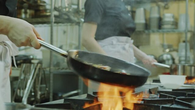 Professional chef tossing food in frying pan while cooking over fire flame above gas stove in the commercial kitchen of restaurant