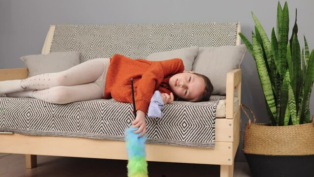 Tired little girl with brown hair being exhausted to help her mother with household chores lying on sofa with pp duster and rag yawning needs to have rest