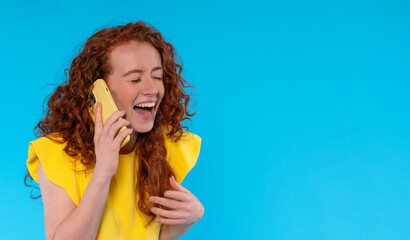 young  laughing redhead positive cute attractive woman in yellow summer dres  holding mobile cell phone chatting on blue color background studio portrait - obrazy, fototapety, plakaty
