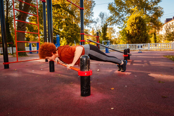 Training in city park. Side view of Caucasian adult woman does push-up from horizontal bar. Concept...