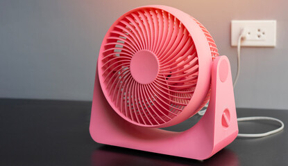 Close up of pink desktop electric fan on black table with soft blurred behind of plug on wall.
