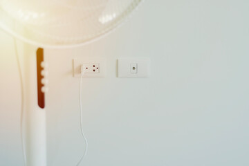 Cropped shot of white modern electric fan in light room with power plug on wall background.