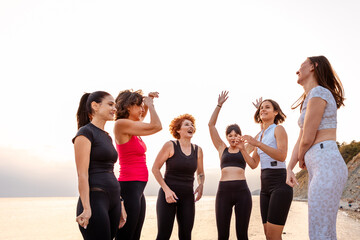 Fototapeta premium Group of adult athletic multiethnic women in sportswear have funny time at coast in background of sunset and sea. Concept of competition and female friendship