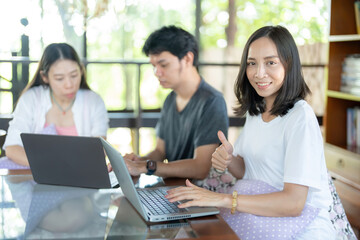 A long-haired Asian female student gives a thumbs up, makes a like sign, uses a laptop to type at work next to two friends working on a project. In the university café there is a library for research.