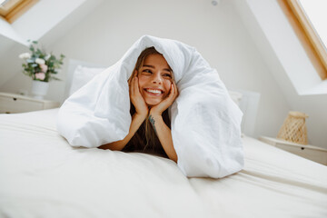 A happy woman is lying on a comfortable bed under a soft blanket and smiling