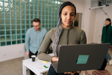 Portrait of African female entrepreneur standing in modern office holding laptop computer, looking to screen. Diverse businesspeople negotiating on blurred background on workplace