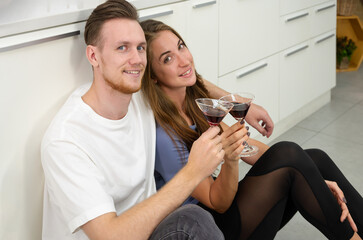 Young couple having fun and drinking cocktails while sitting on kitchen floor at home and looking at camera. - 768587149