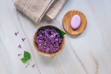 Spa and wellness composition with perfumed lilac flowers water in wooden bowl, terry towels and handmade soap on marble background. Aromatherapy, top view, flat lay, copy space.