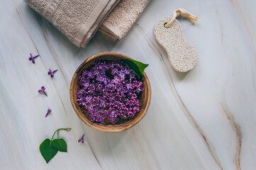 Lilac spa. Towels, pumice stone for feet, lilac flowers. Natural herbal cosmetics with flowers on marble background. Relaxation concept. Cosmetic procedures. Copy space.
