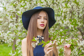 Elegant brunette woman portrait. Beautiful female model with long hair and make-up wearing classic blue hat in spring garden outdoor - 768591371