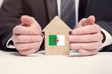 Businessman hand holding wooden home model with Algerian flag. insurance and property concepts
