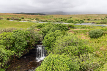 Fototapeta na wymiar Nestled in lush greenery, the Lealt Falls create a tranquil haven on Isle of Skye, complemented by a countryside road and the distant, fog-shrouded highlands