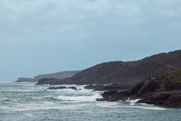Fototapeta premium A moody view of Scourie Bay, where tumultuous waves crash against rugged cliffs under a somber sky. The coastline, a blend of raw beauty and wild nature