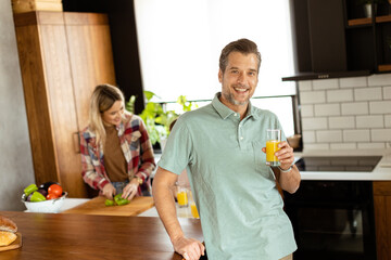 Smiling man holds a glass of orange juice while two women cook in a bright kitchen - 768597773