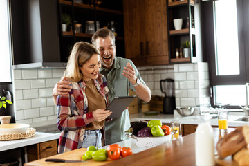 Cheerful couple stands in a well-lit kitchen, engrossed in a digital tablet among fresh ingredients - 768597949