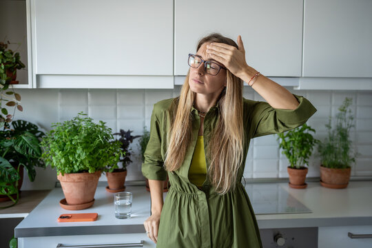 Tired exhausted blonde woman feels headache touching head with closed eyes on kitchen at home. Unhealthy female in glasses suffers head pain, dizziness, migraine, cephalalgia feeling unwell pressure.