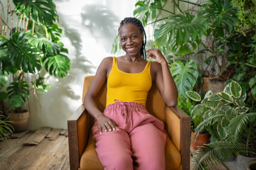 Happy African American woman relaxing on chair in cozy green apartment with monstera plants, cheerful black girl spending time, looking at camera, resting in armchair at home garden. Mindfulness. 