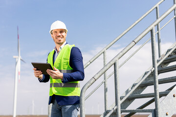 Smiling Engineer With Tablet Inspecting Wind Turbines On A Sunny Day - 768598938