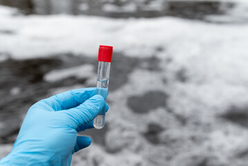 Fototapeta premium Test tube with red cap and water inside in male hand in blue medical gloves, close up. Foaming dirty water on the background, concept of water pollution