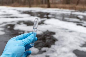 Male hand in blue medical gloves holding test tube with water inside, close up. Checking the pollution level of foaming water in unclean river on the background