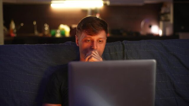 Relaxed young man vaping e-cigarette sitting on sofa while watching movie or sporty match on laptop at home in evening after work. Handsome male smoking ecigarette enjoy moment, looking to screen.