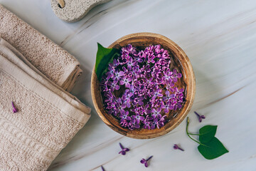 Spa and wellness composition with perfumed lilac flowers water in wooden bowl and terry towels on marble background, aromatherapy, top view, flat lay.